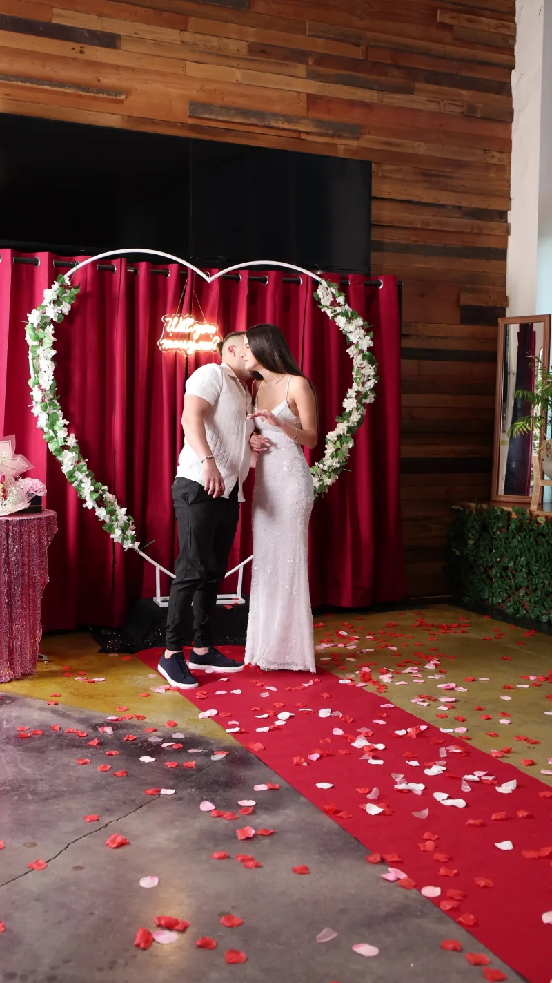 A couple under a white flower heart arch with a neon sign and rose petals on the floor in the Main Hall at 1712 Studios, front windows behind
